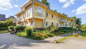 A multi-story yellow building with balconies, surrounded by greenery and a paved driveway.