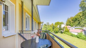 A balcony with a table and chairs, a vase with flowers, and a view of a green lawn and houses.