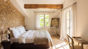 Une chambre avec un mur en pierre, des poutres en bois, un lit avec du linge de maison blanc, un tableau posé sur une petite table et une fenêtre avec des rideaux.