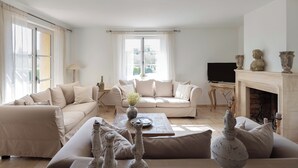 Un salon spacieux avec un ensemble de canapés beige, une table basse agrémentée d’objets décoratifs, une cheminée et un téléviseur.