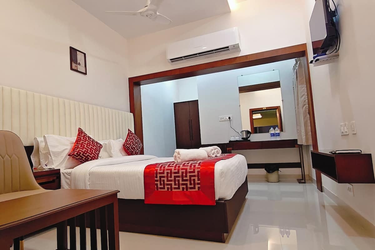 A hotel room with a bed, two chairs, a small table, a mirror, and an air conditioning unit.