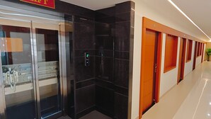 A hotel corridor with a glass elevator, a reception area, and a seating area with a plant. - Aswin Residency (Namakkal)
