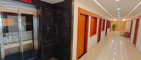 A hotel corridor with a glass elevator, a reception area, and a seating area with a plant.