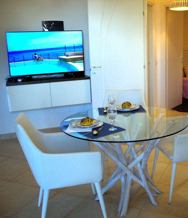 Uma área de refeições moderna com uma mesa de vidro, cadeiras brancas e uma TV de tela plana exibindo uma paisagem de praia.