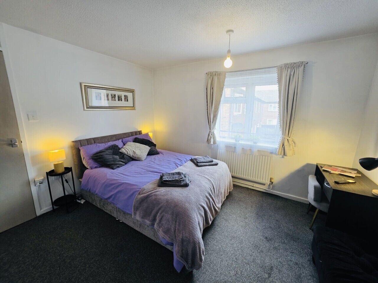 Un dormitorio con cama, mesita de noche, lámpara y una ventana con cortinas.