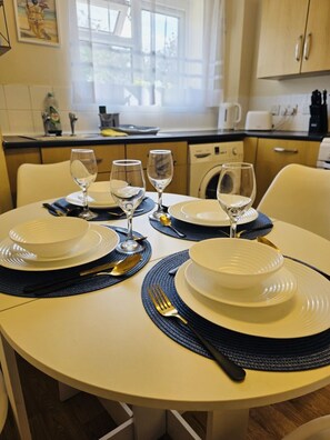 A dining table set with white plates, bowls, and cutlery, with a kitchen in the background. - The Nook perfect for families, corporate and business travelers (West Northamptonshire)
