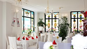 A dining area with white tablecloths, red napkins, and chairs with white covers. There are potted plants and a chandelier. - Ratskeller Hotel & Restaurant Lindow  (Lindow)