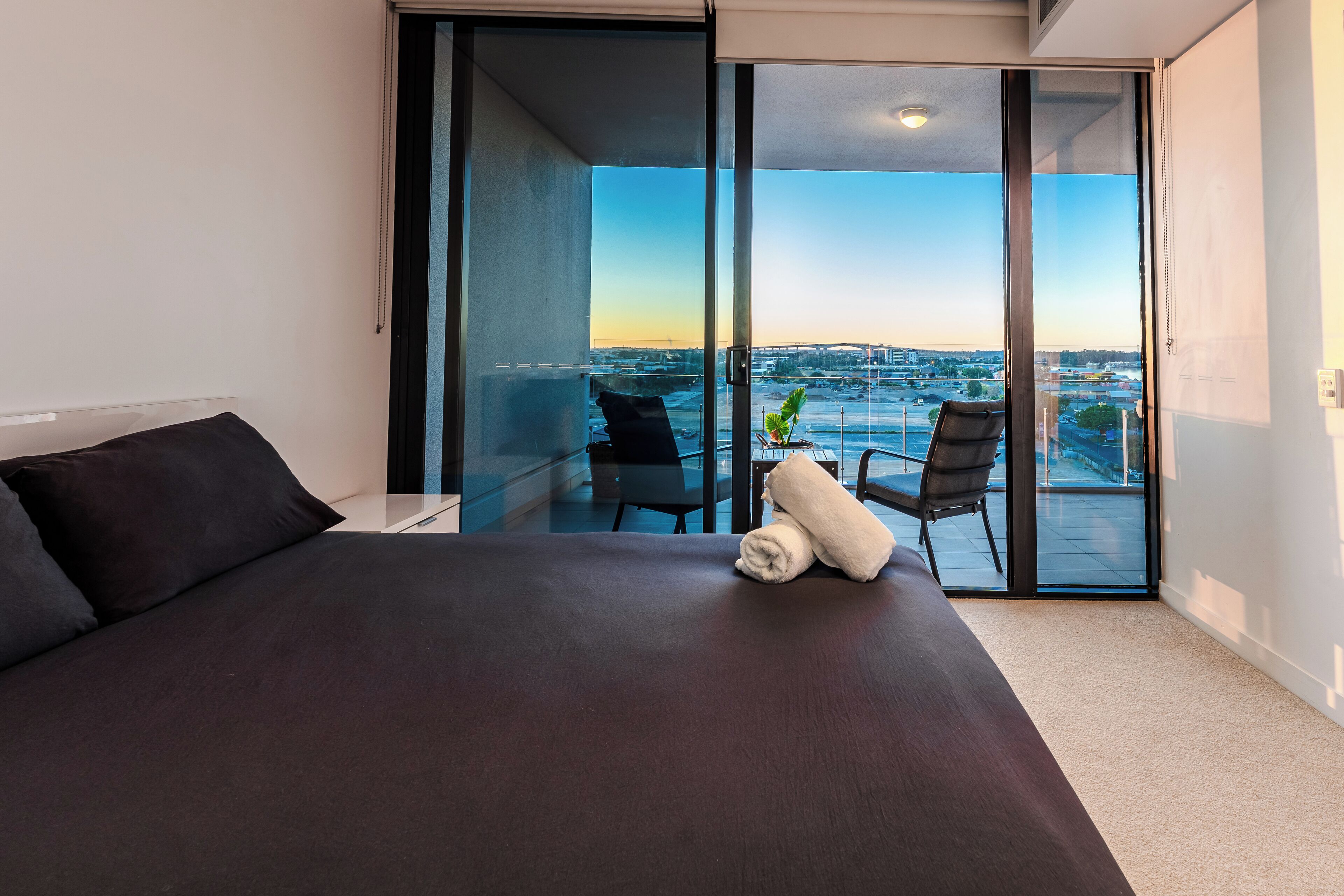Et moderne soveværelse med en stor seng, udsigt til byen gennem gulv til loft-vinduer og en balkon med stole.