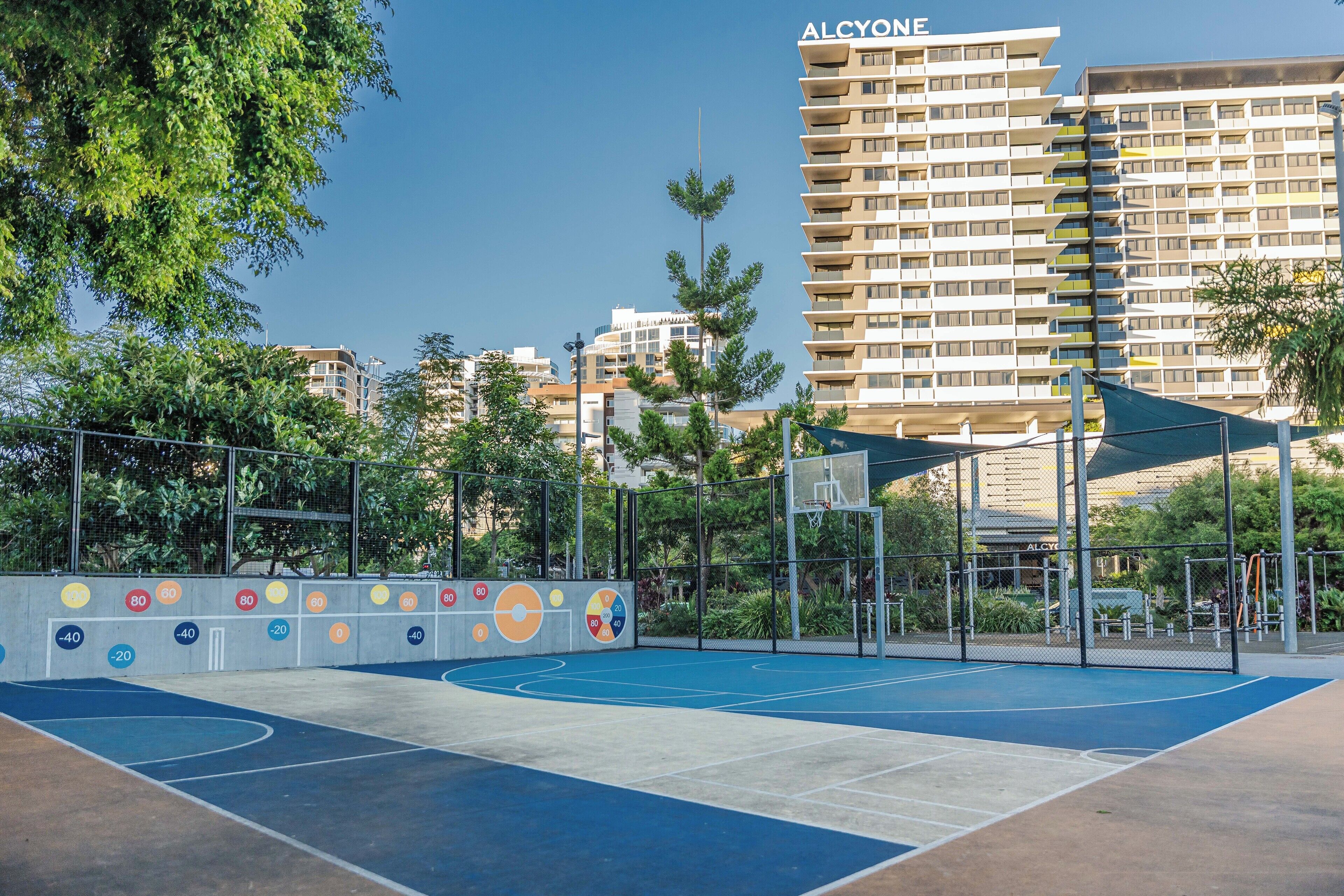 An outdoor basketball court with a net, surrounded by a fence and trees. In the background, there is a tall building with the name "ALCYONE" visible.