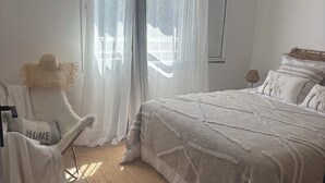 Una camera da letto con un letto, una sedia, una finestra con tende, una ventola a soffitto e un tappeto intrecciato.