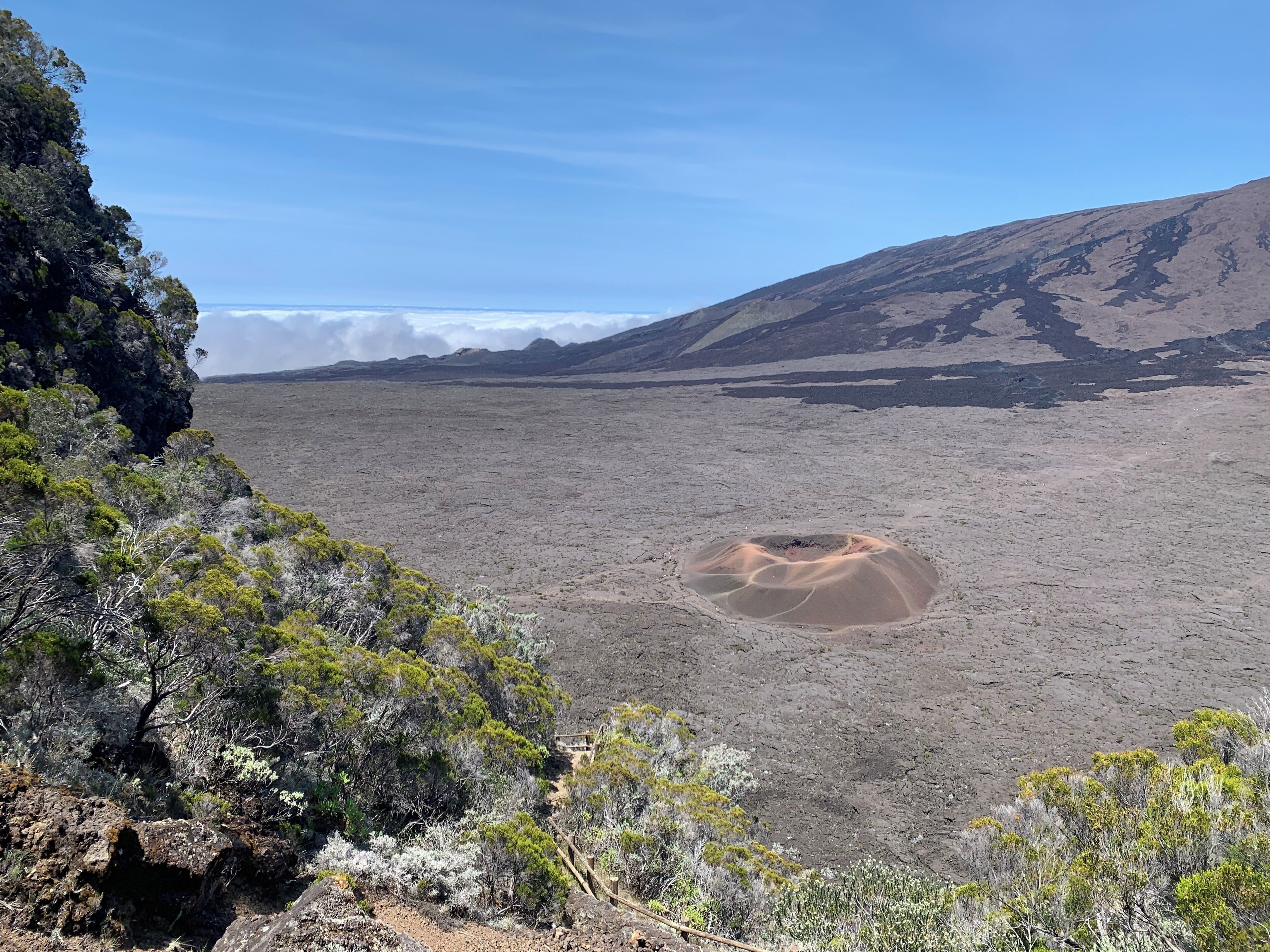 A barren landscape with sparse vegetation, a crater, and a distant mountain range under a clear sky.
