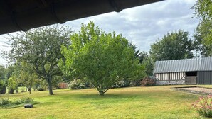 A covered outdoor area with a wooden deck, a metal table, and a grassy lawn with trees and a building in the background. - Gîte near Honfleur (Gonneville-sur-Honfleur)