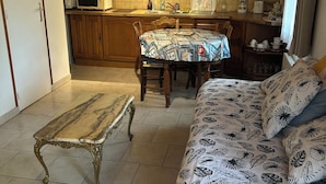 A cozy living room with a sofa, a small table, and a dining table with chairs. A kitchen is visible in the background with a microwave and sink. - Gîte near Honfleur (Gonneville-sur-Honfleur)