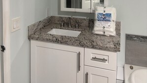 Ein Badezimmer mit einem weißen Waschbecken, einer Granit-Arbeitsplatte und einem Spiegel darüber. Drei Leuchten sind über dem Spiegel angebracht, und auf der Arbeitsplatte liegt eine Packung Handtücher.