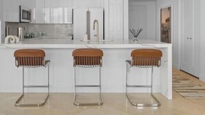 Eine moderne Küche mit weißen Schränken, einer zentralen Kochinsel mit Hockern, einem Edelstahlkühlschrank und einer Pendelleuchte.