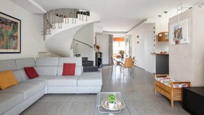 Una sala de estar moderna con un sofá blanco, una mesa de centro con una planta, una escalera de caracol y un comedor al fondo.