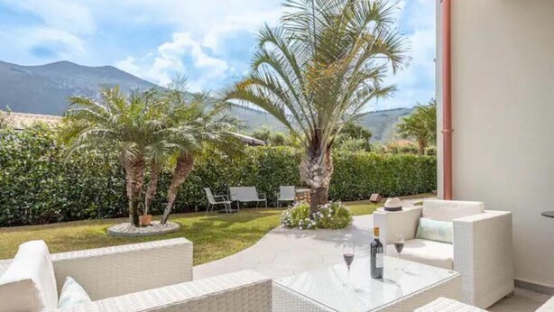 Un patio con mobiliario de ratán blanco, una mesa de vidrio con una botella y un vaso, y vista a una montaña y palmeras.