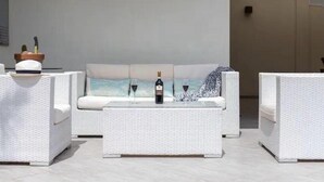 Un sofá blanco de mimbre con un cojín, una mesa de centro con superficie de vidrio y dos sillones blancos. Hay una botella de vino y dos copas de vino sobre la mesa.