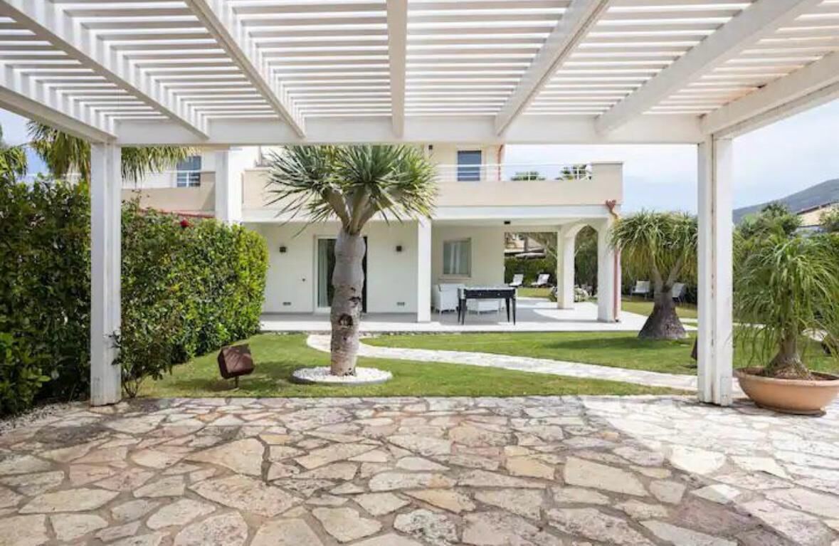 A covered outdoor area with stone flooring, a palm tree, and a building in the background.
