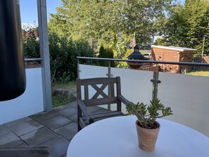 Ein Balkon mit einem Holzstuhl, einem Blumentopf und Blick auf ein Holzgebäude sowie Bäume.