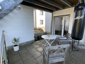 Eine Terrasse mit einem runden Tisch, zwei Holzstühlen, einem Sandsack und einem Blumentopf.
