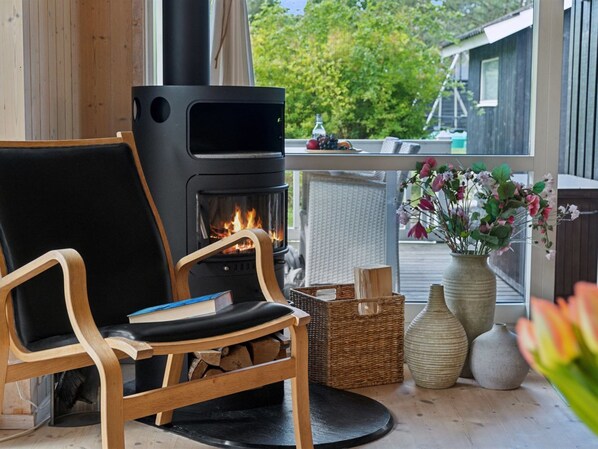 Une pièce chaleureuse avec un poêle à bois, un fauteuil confortable, une petite table et une vue sur l’extérieur.