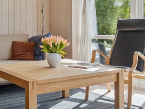 Una mesa de madera con un florero, una revista y una silla con cojín negro en una habitación con paredes de madera y una ventana.