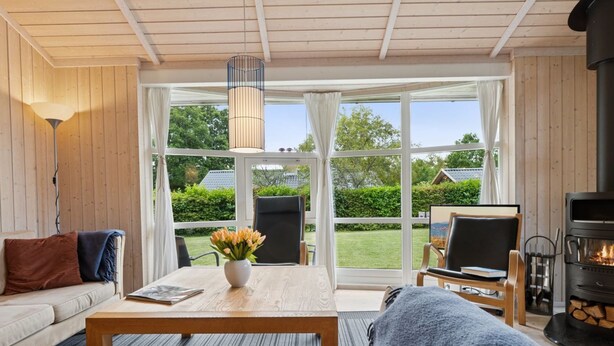 Ein gemütliches Wohnzimmer mit einer Couch, einem Couchtisch mit Blumenvase, einem Kamin und großen Fenstern mit Blick ins Freie.