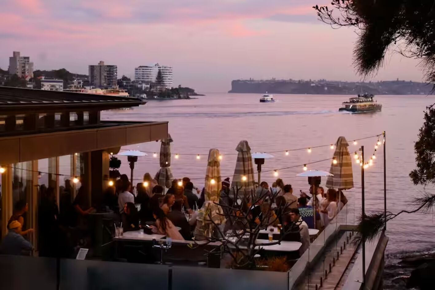 A waterfront restaurant with outdoor seating, string lights, and umbrellas.
