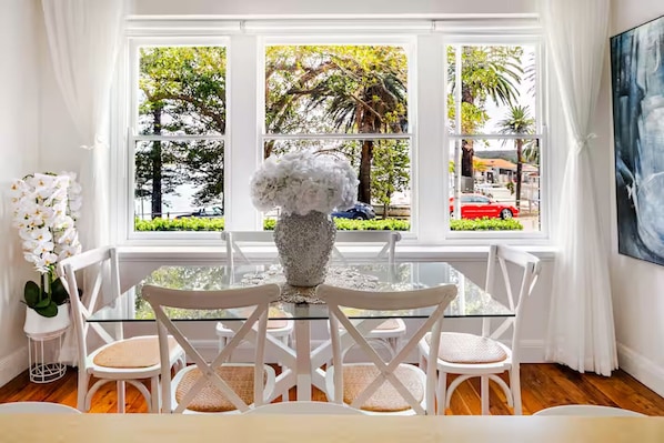 A dining area with a glass table, white chairs, a vase with white flowers, and a view of trees and a red car through large windows.