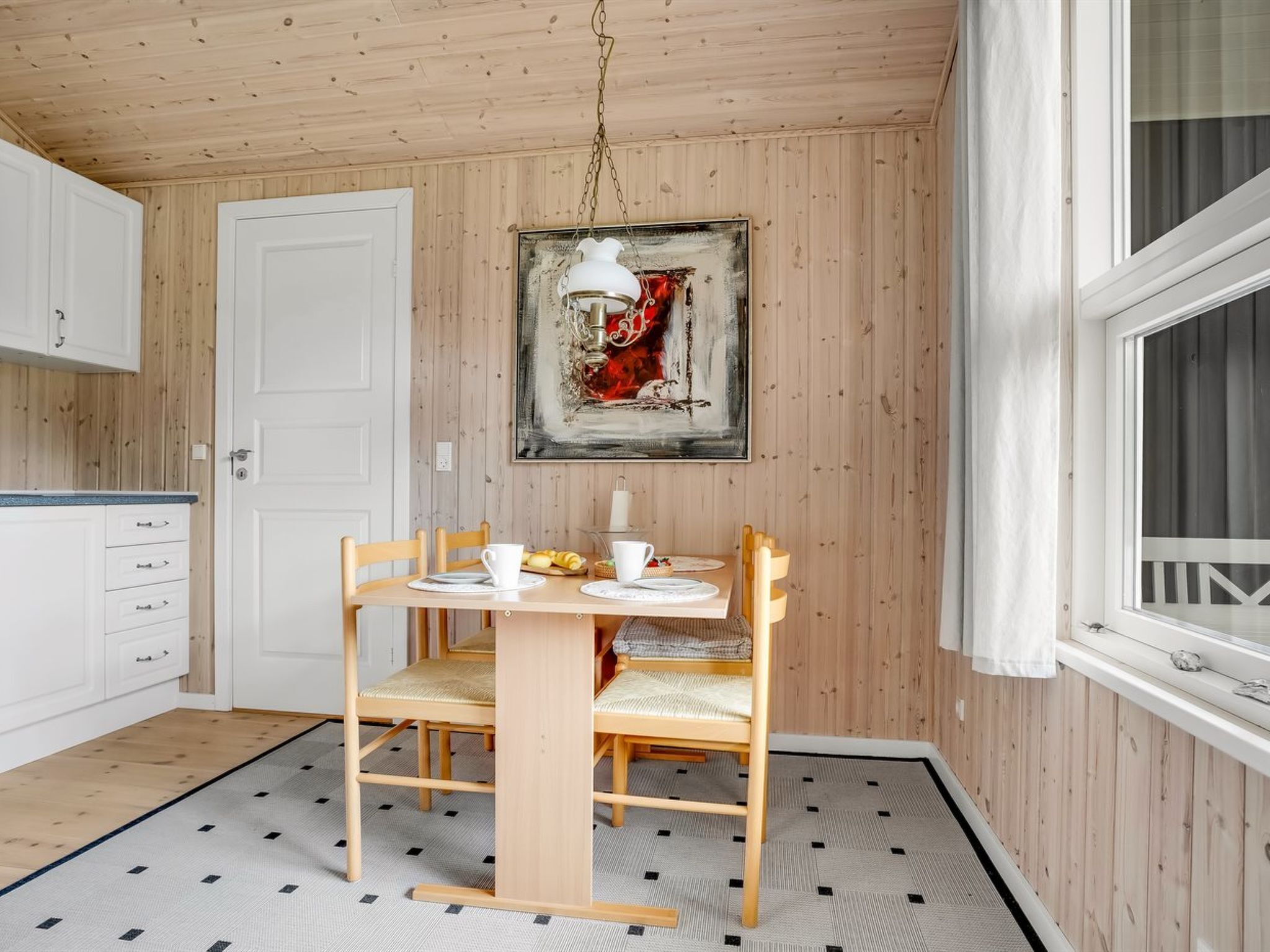 A compact kitchen with wooden cabinets, a dining table with chairs, a hanging light fixture, and a large window with curtains.