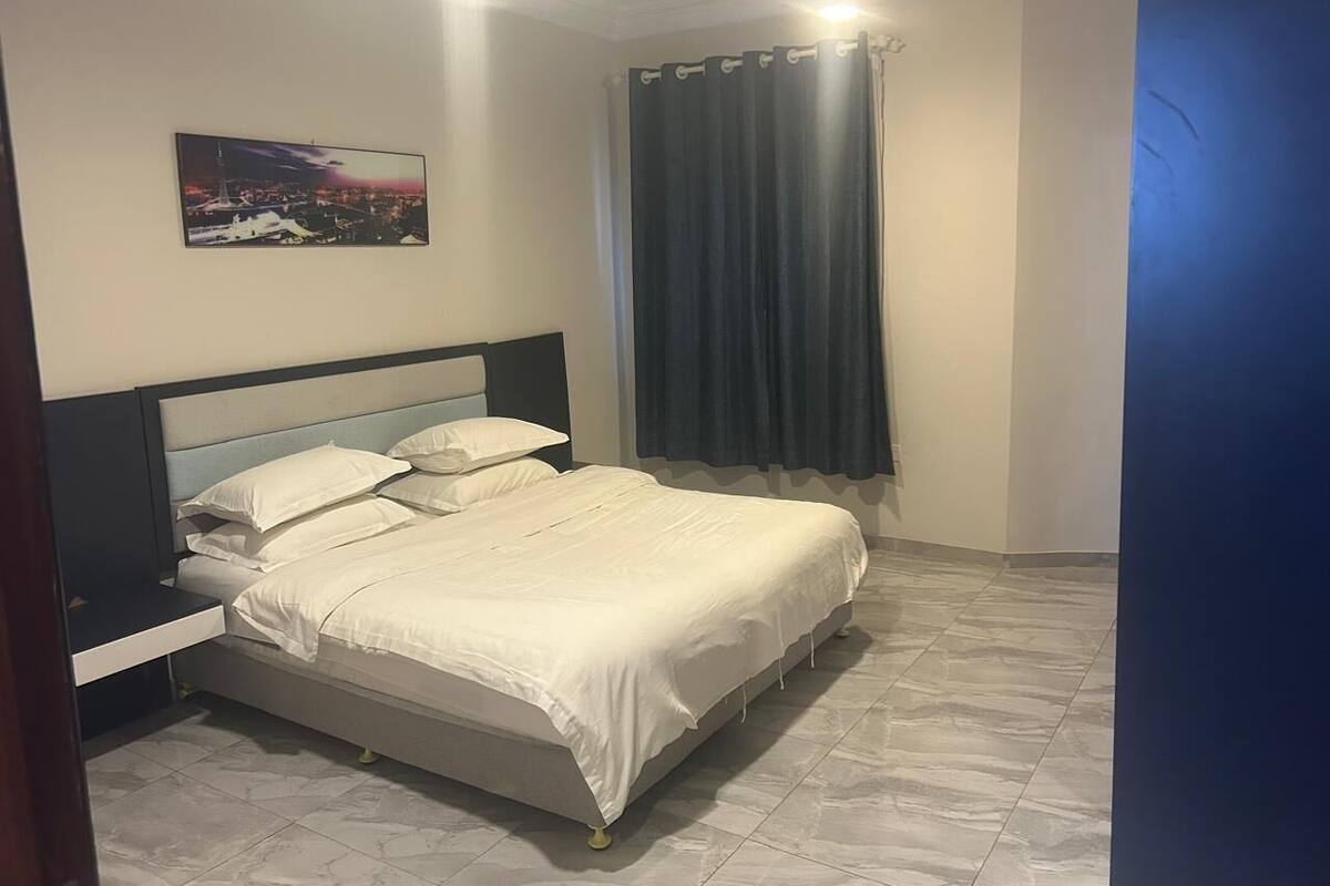 A neatly made bed with white bedding, a nightstand, a wall-mounted picture, and a window with dark curtains.