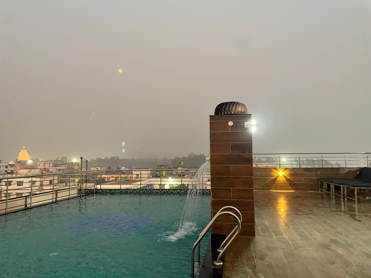 A rooftop swimming pool with a water fountain, a view of the cityscape, and a bench area.