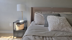 Una camera da letto ordinata con un letto, un comodino, una lampada e un cesto.