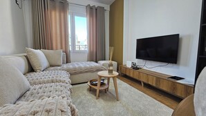 Una sala de estar moderna con un sofá, un reposapiés y una mesita con un objeto decorativo. Un televisor de pantalla plana está montado en la pared, y se ve una ventana con cortinas.