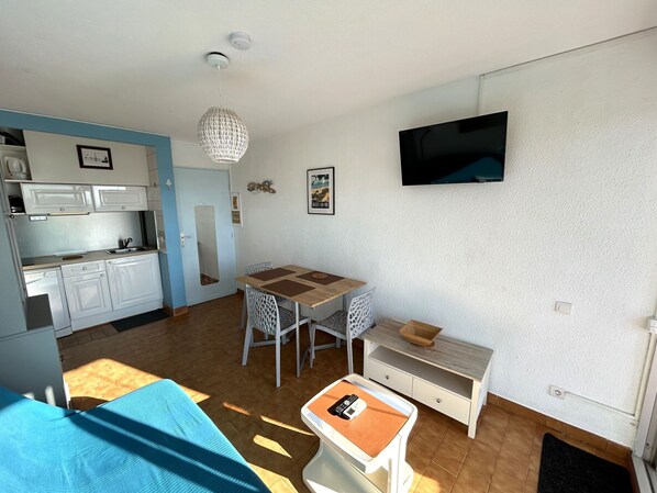 A compact living space with a kitchenette, a dining area, a TV mounted on the wall, and a small table with a chair.