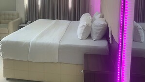 Una cama bien hecha con ropa blanca y almohadas, una mesita de noche con un teléfono y un espejo grande con luces LED rosas.