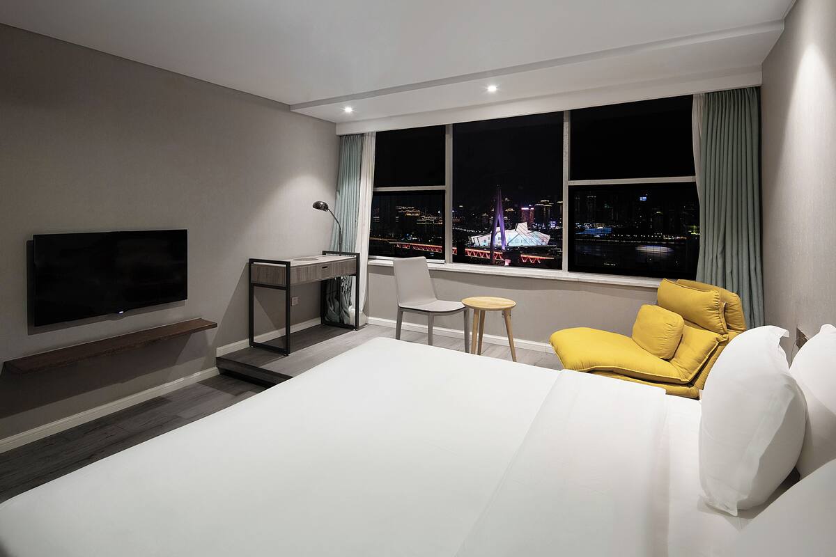 Une chambre d’hôtel équipée d’un lit, d’un bureau, d’une chaise, d’une télévision et d’une grande fenêtre donnant sur la ville.