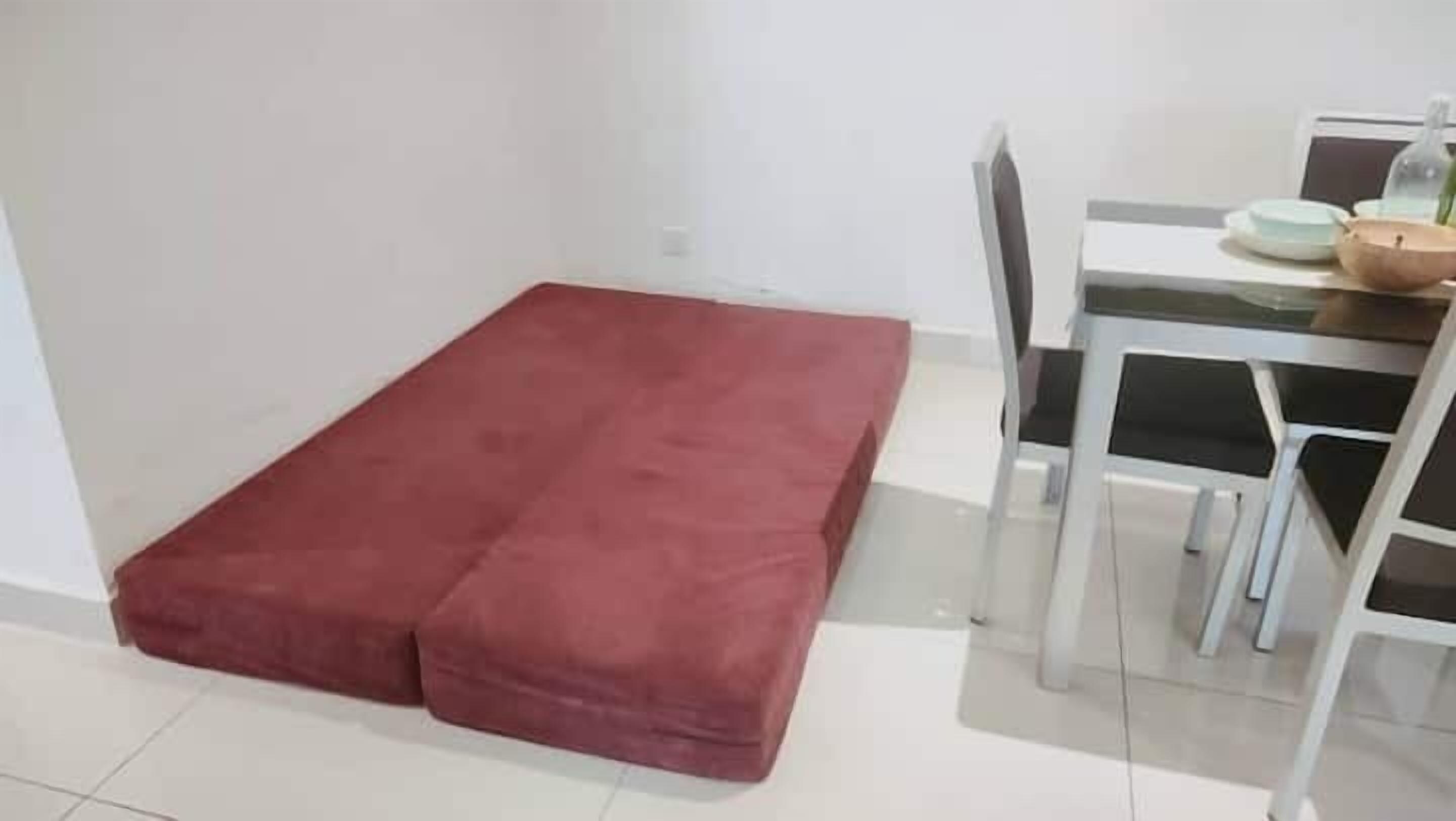 A room with a red mattress on the floor, a white table with chairs, and a small table with a bowl and a plant.