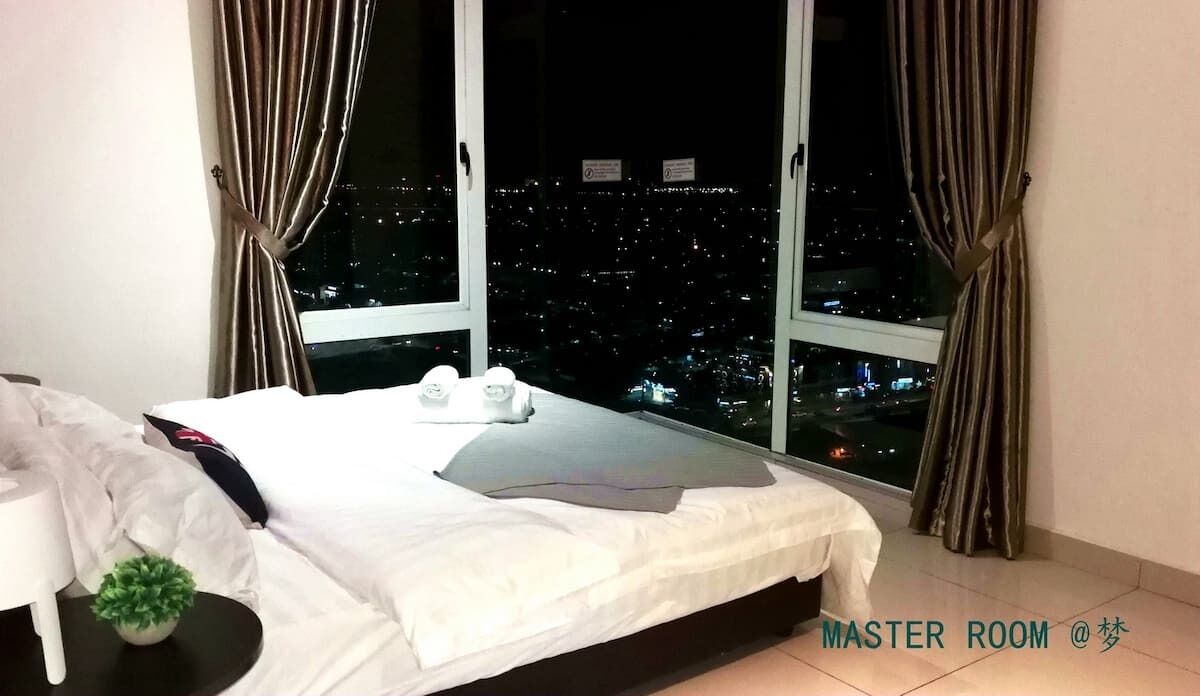 Une chambre principale avec un grand lit, une petite table de chevet, une plante en pot et une vue sur le paysage urbain illuminé la nuit.
