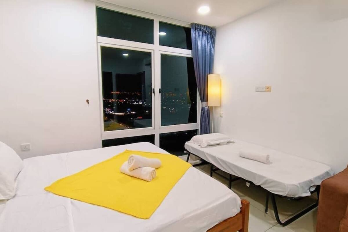 A hotel room with a bed, a massage table, a window with a view of the city at night, and a chair.
