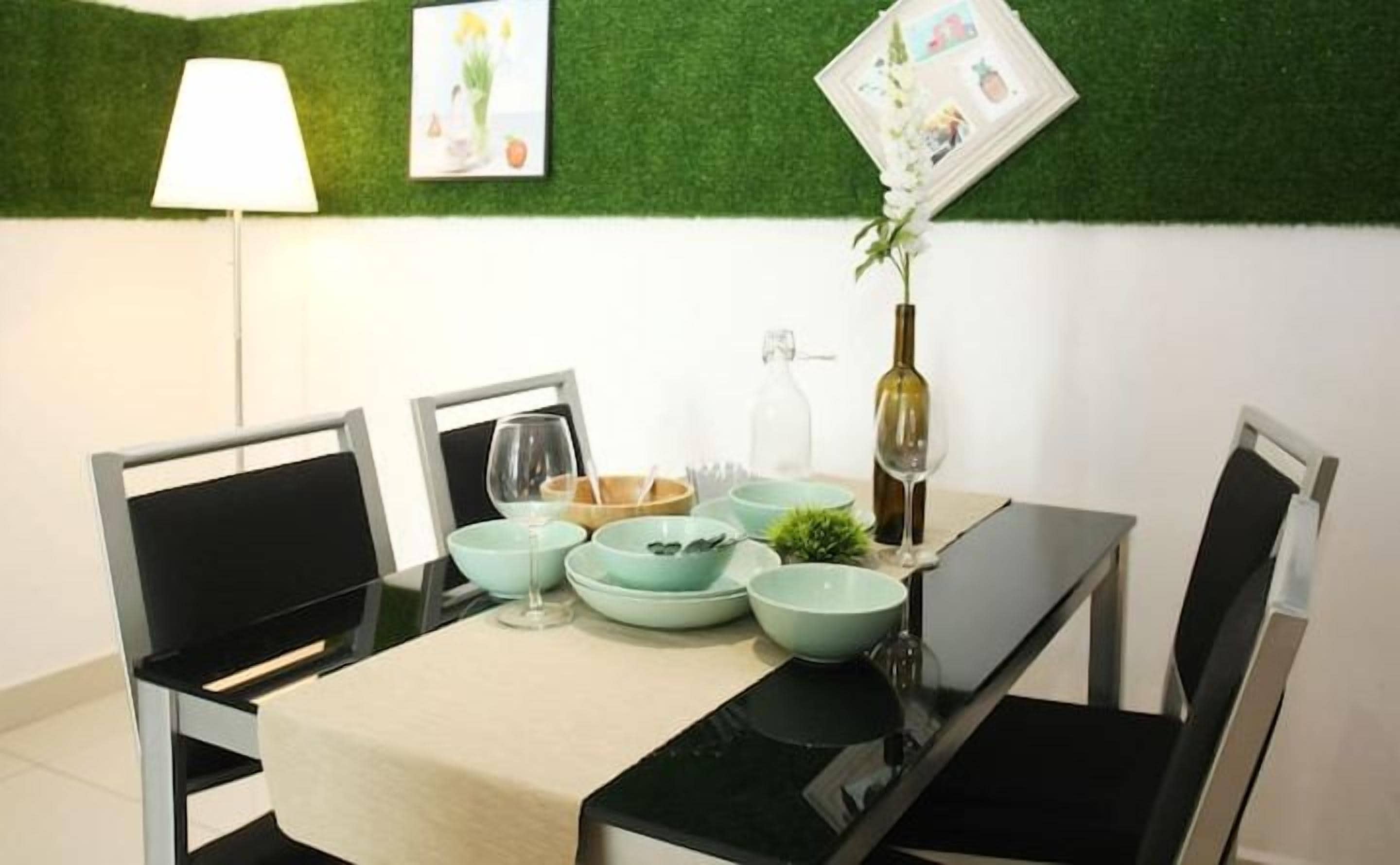 A dining area with a table set for four, chairs, a lamp, and a green wall with a framed picture.