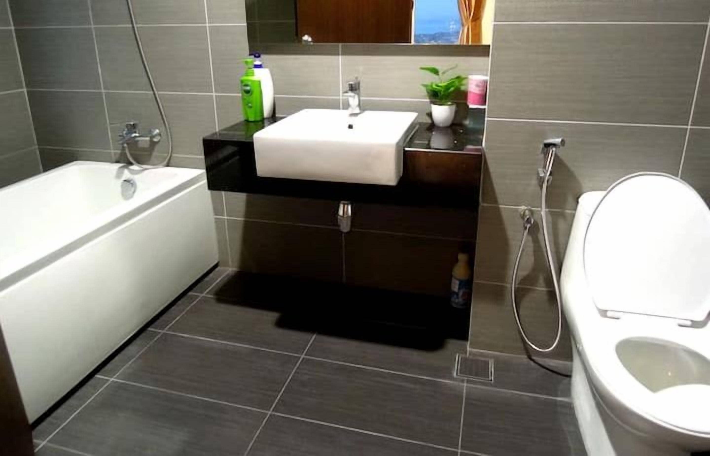 Ein modernes Badezimmer mit einer Badewanne, einem Waschbecken mit Spiegel darüber und einer Toilette. Auf der Ablagefläche des Waschbeckens steht eine kleine Pflanze, und im Hintergrund ist ein Fenster mit Vorhängen zu sehen.