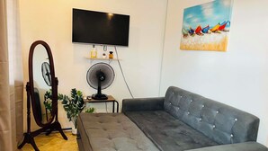 Een woonkamer met een grijze bank, een vloerventilator, een flatscreen televisie, een schilderij van boten en een klein tafeltje met decoratieve objecten.