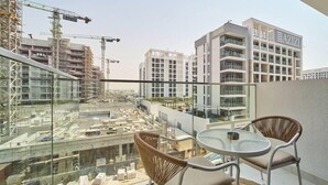 A balcony with two wicker chairs and a glass table, overlooking a construction site and a building with the sign "AZIZI".