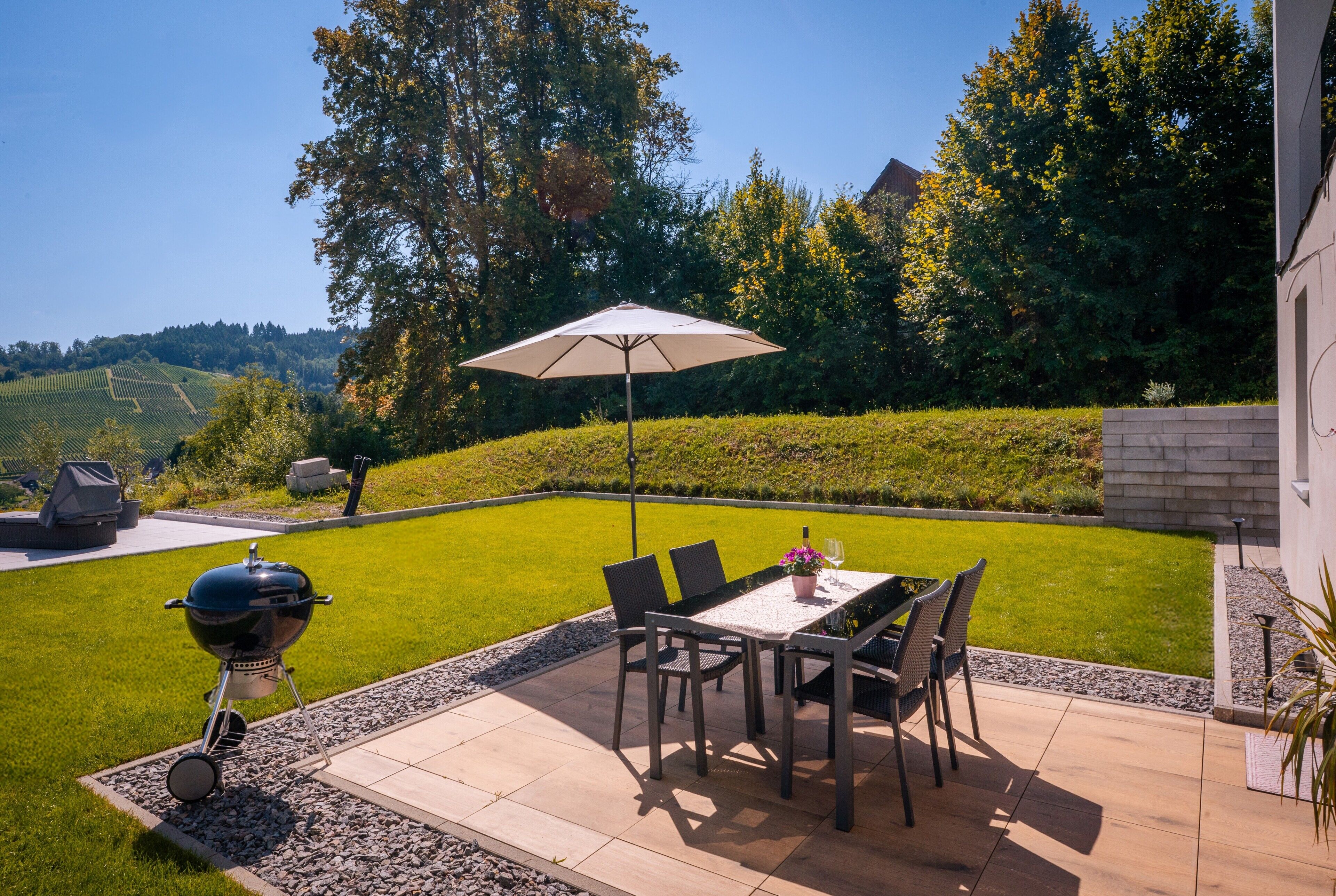 Eine Terrasse mit Tisch, Stühlen, einem Grill und Blick auf einen angelegten Garten und Hügel.
