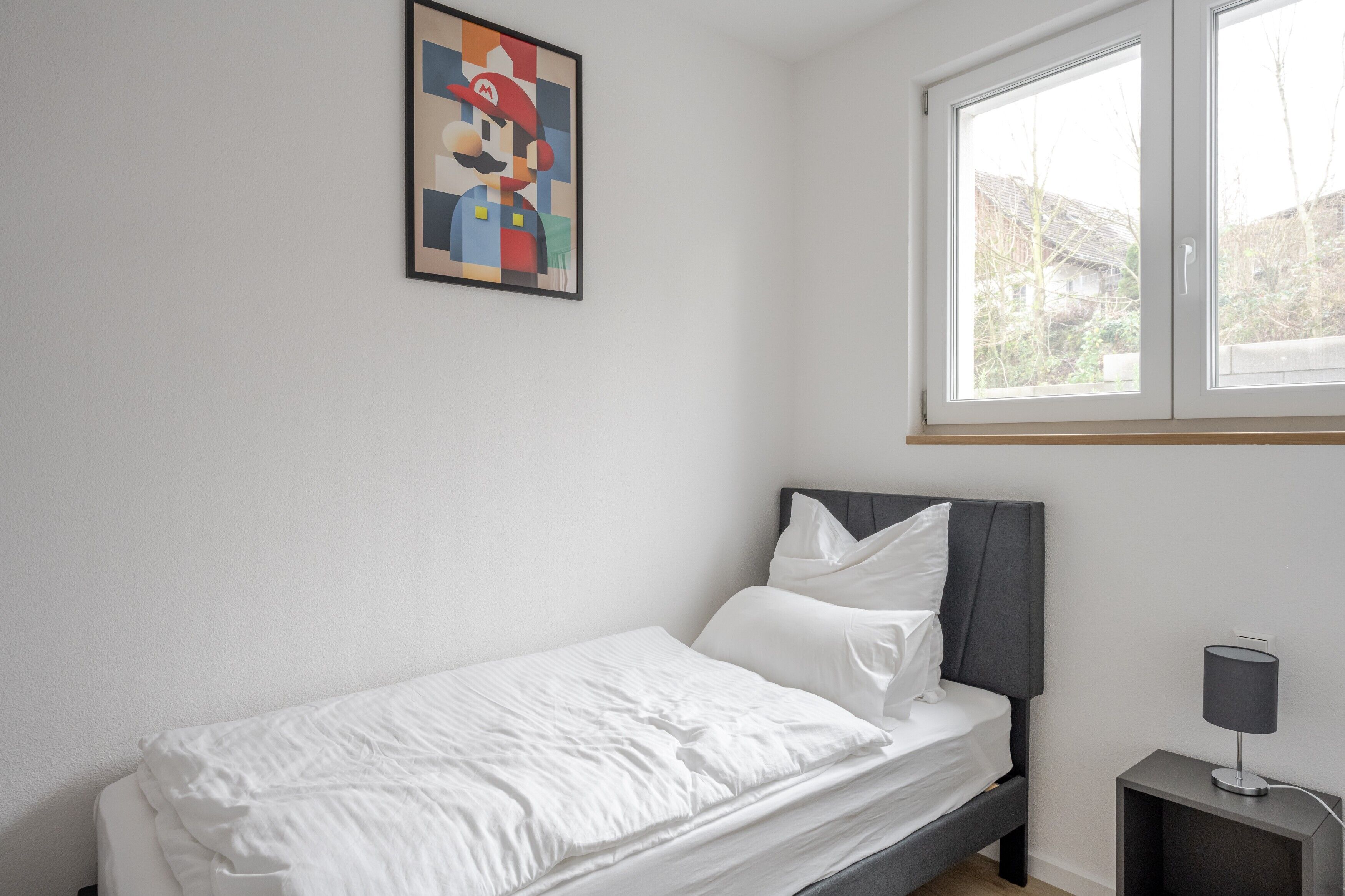 A single bed with white bedding, a gray headboard, a bedside table with a lamp, and a framed picture on the wall.