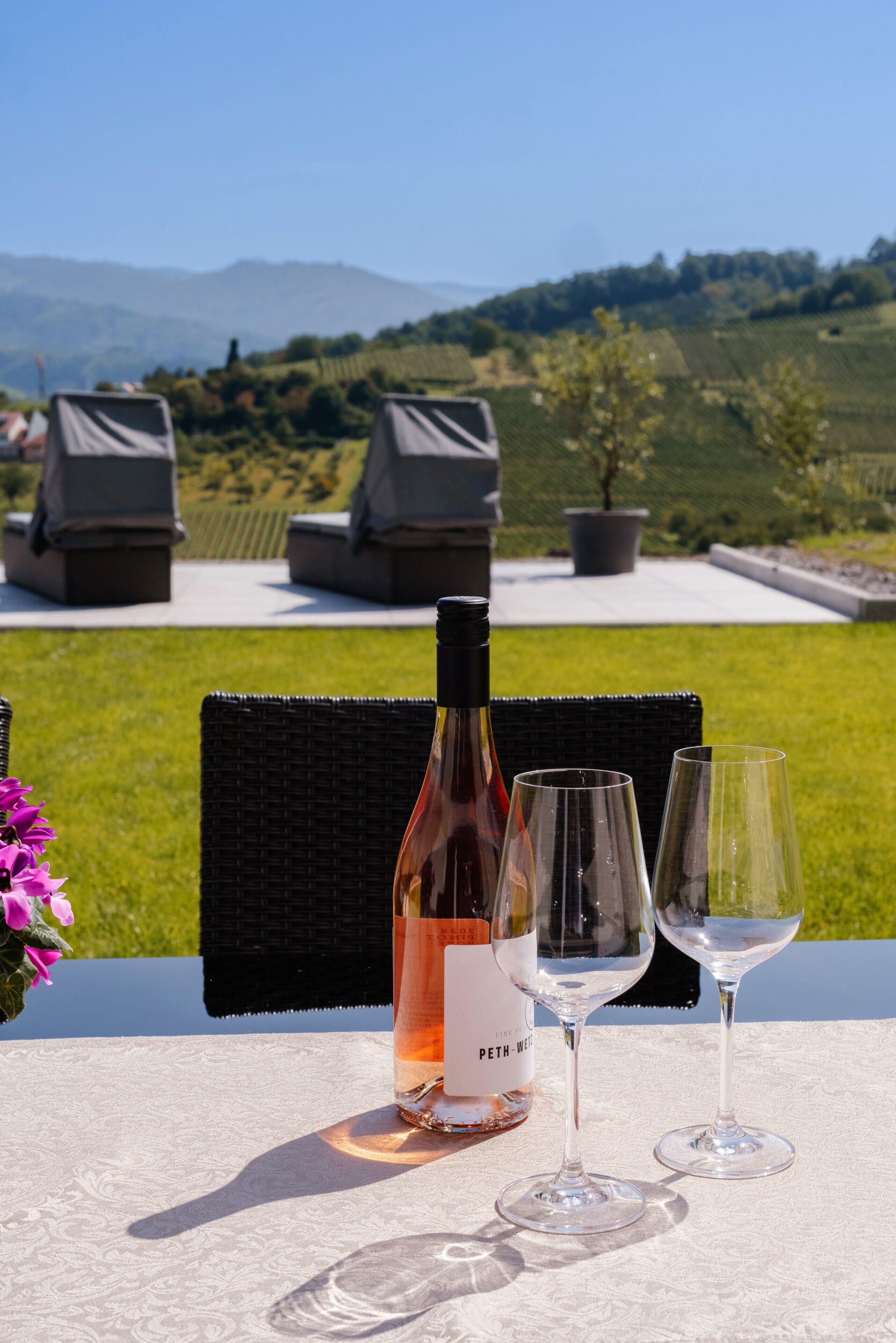 A table with a bottle of wine and two glasses, set outdoors with a view of a vineyard and hills in the background.
