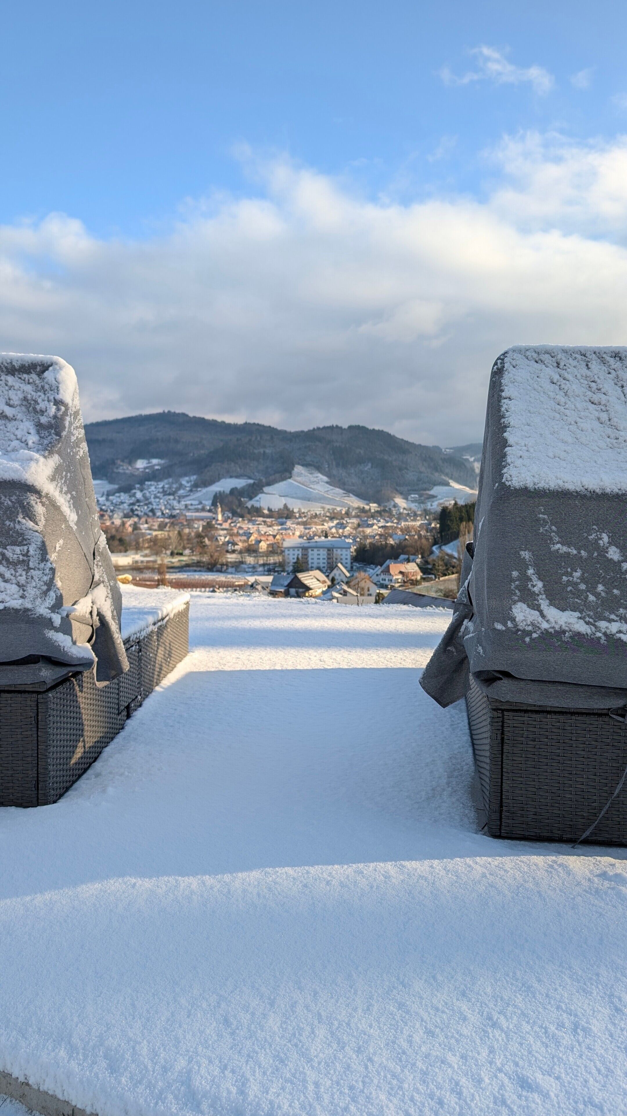 A snow-covered outdoor seating area with wicker chairs, a clear view of a town in the distance, and a mountain range in the background.