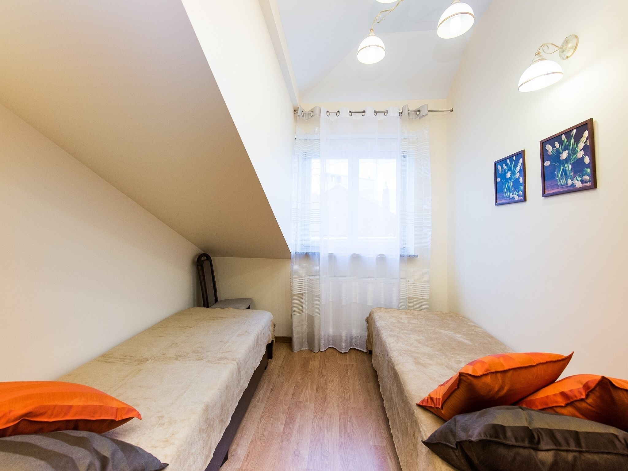 Una habitación pequeña y luminosa con dos camas individuales, suelo de madera, una ventana con cortinas translúcidas y dos cuadros enmarcados en la pared.